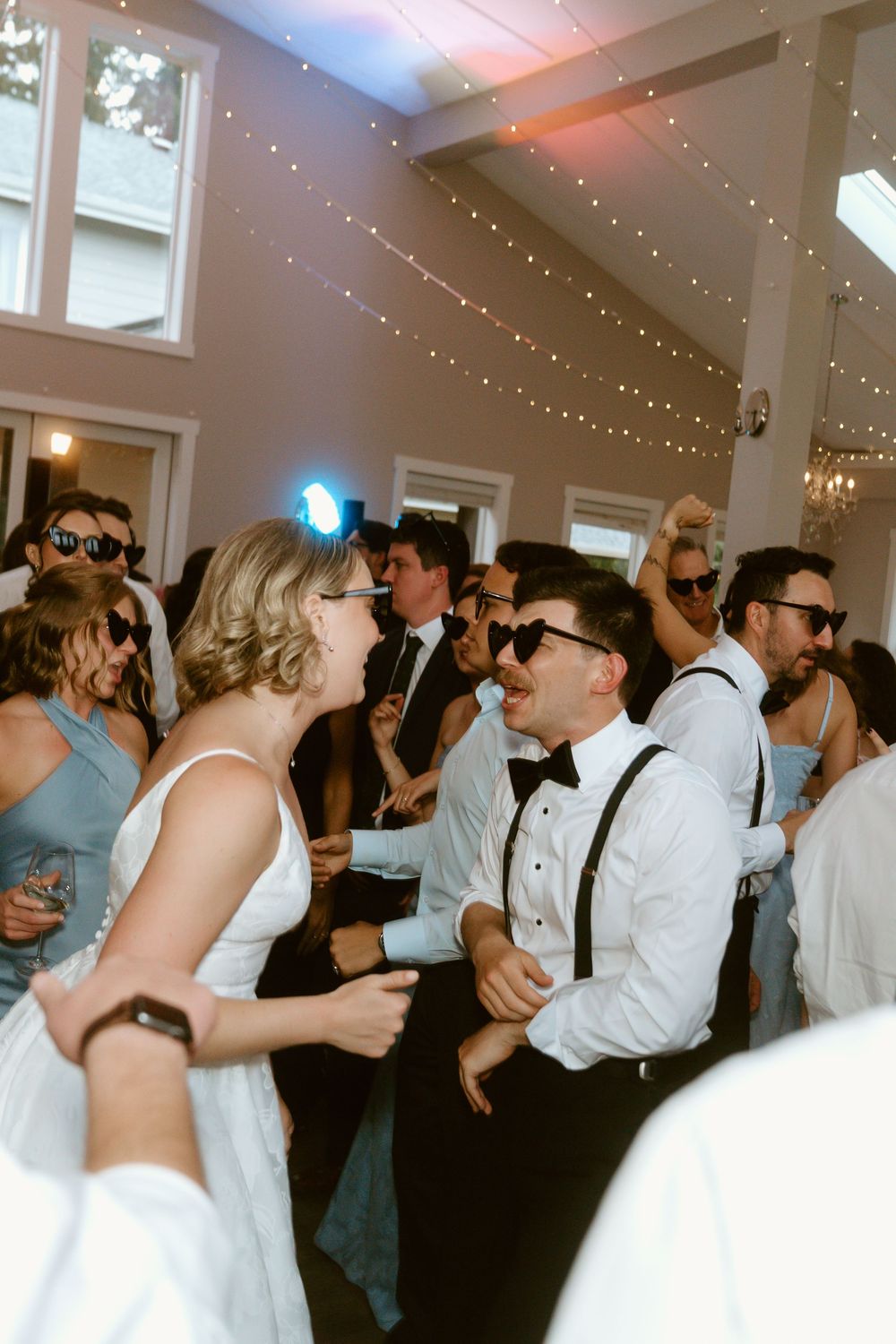 Bride and groom dancing joyfully with guests at a wedding reception, wearing sunglasses under twinkling fairy lights.