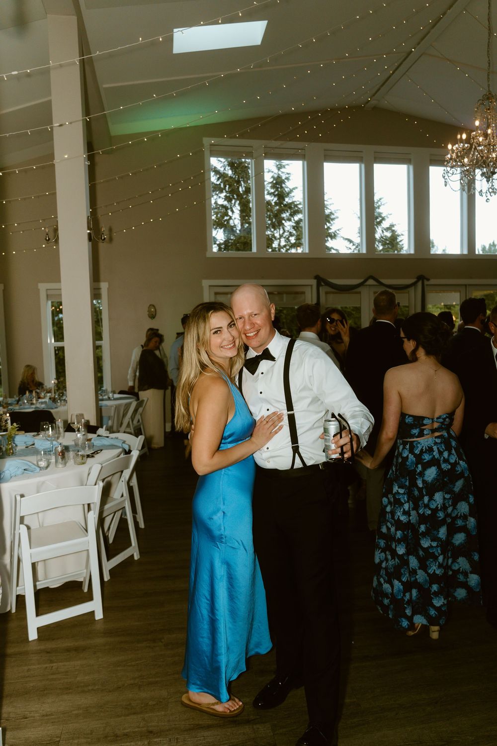 A couple poses together at a wedding reception, woman in blue gown and man in tuxedo with suspenders, fairy lights overhead.