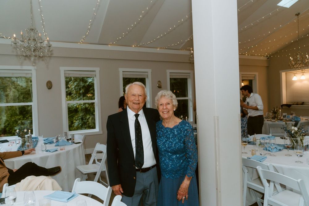 Elderly couple smiling together at an elegant wedding reception venue with white chairs and blue table settings.