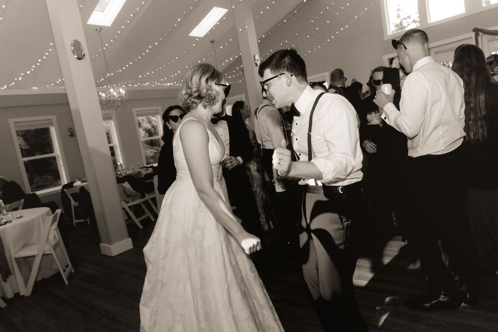 Black and white photo of bride and groom dancing joyfully at their wedding reception with guests in background.