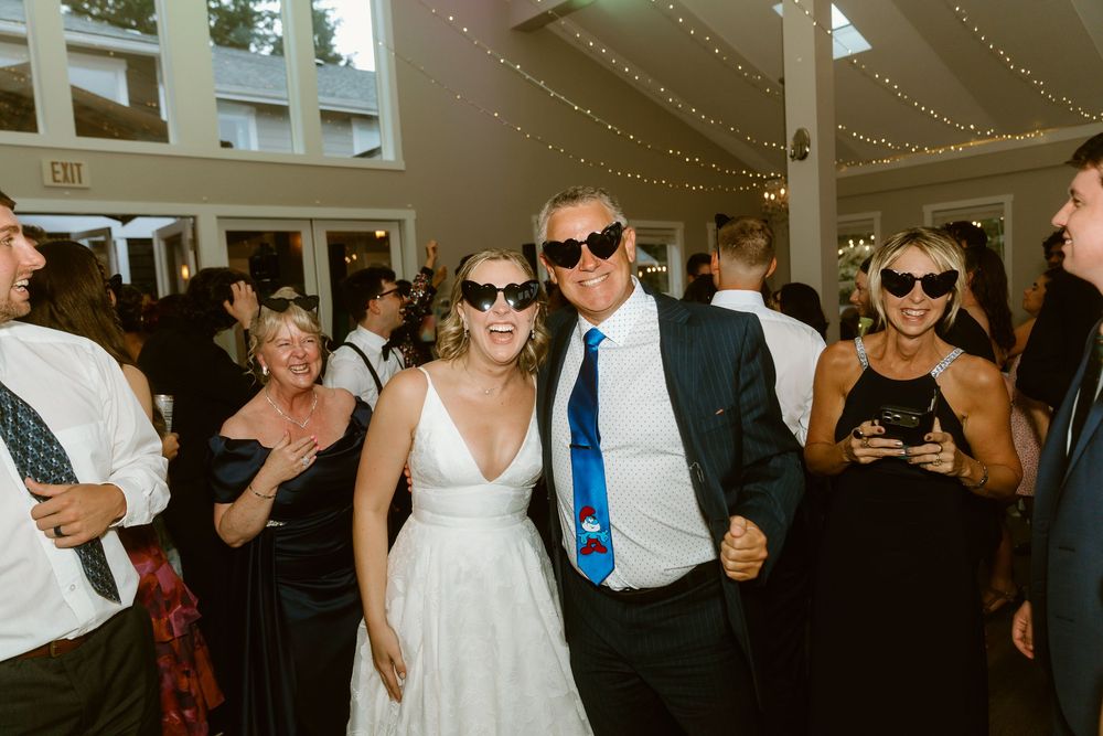 Bride and groom wearing sunglasses dancing joyfully with guests at an elegant indoor wedding reception.