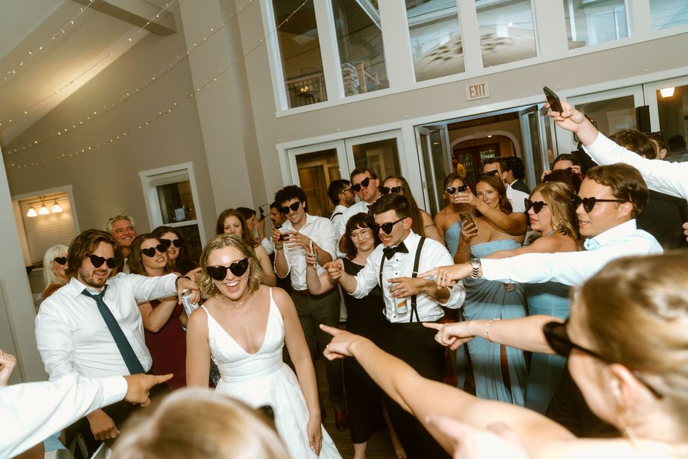 Wedding reception crowd wearing sunglasses celebrating with bride and groom on dance floor indoors.