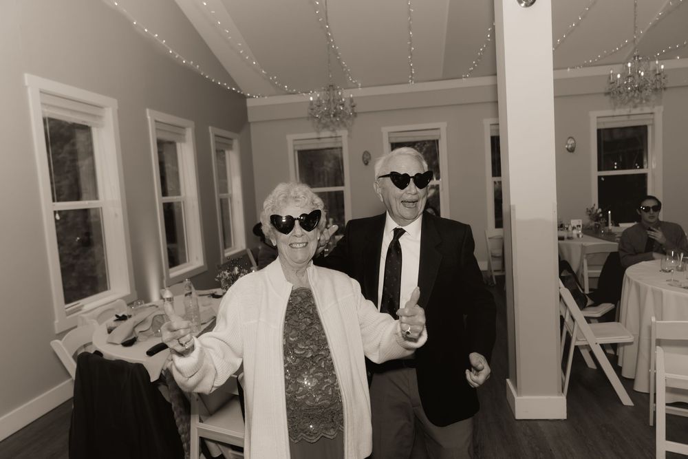 Two elderly people wearing sunglasses dancing joyfully at an indoor reception venue in black and white photo.
