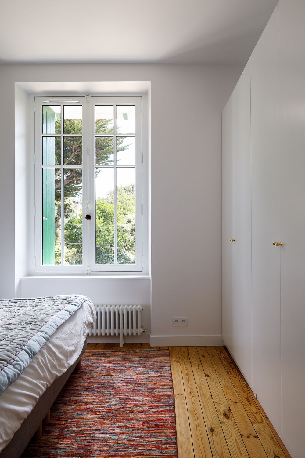 Chambre blanche minimaliste avec parquet, tapis rouge, placard intégré et fenêtre donnant sur le jardin.