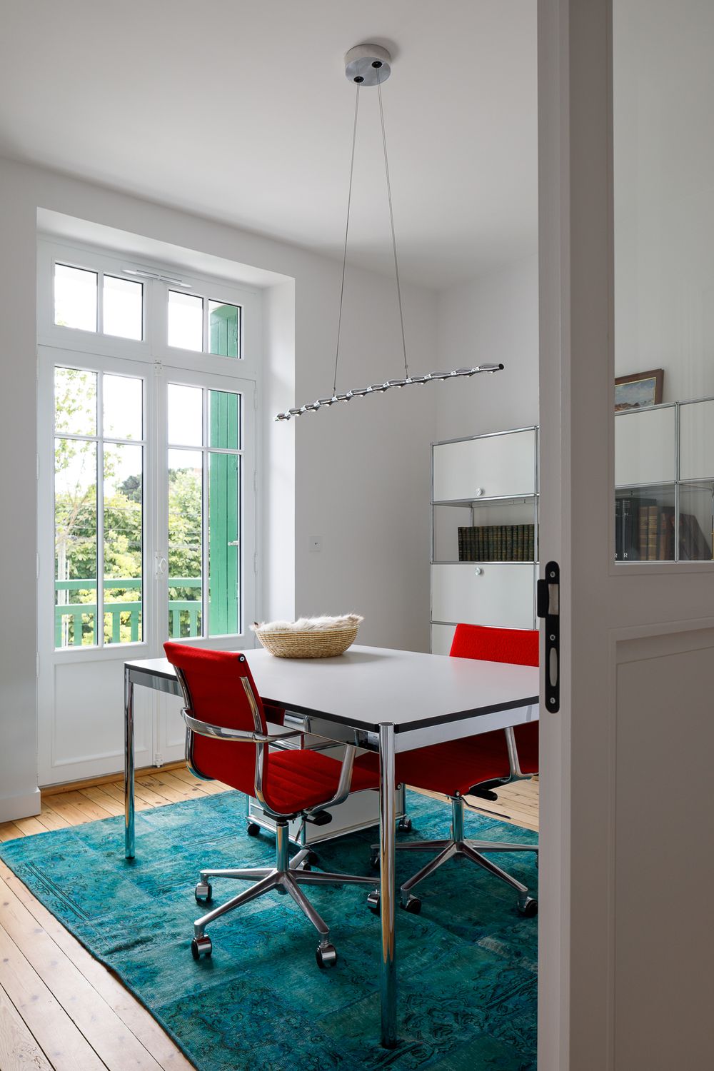 Bureau avec des chaises rouges, une table blanche, un tapis bleu sarcelle et une grande fenêtre donnant sur le jardin.
