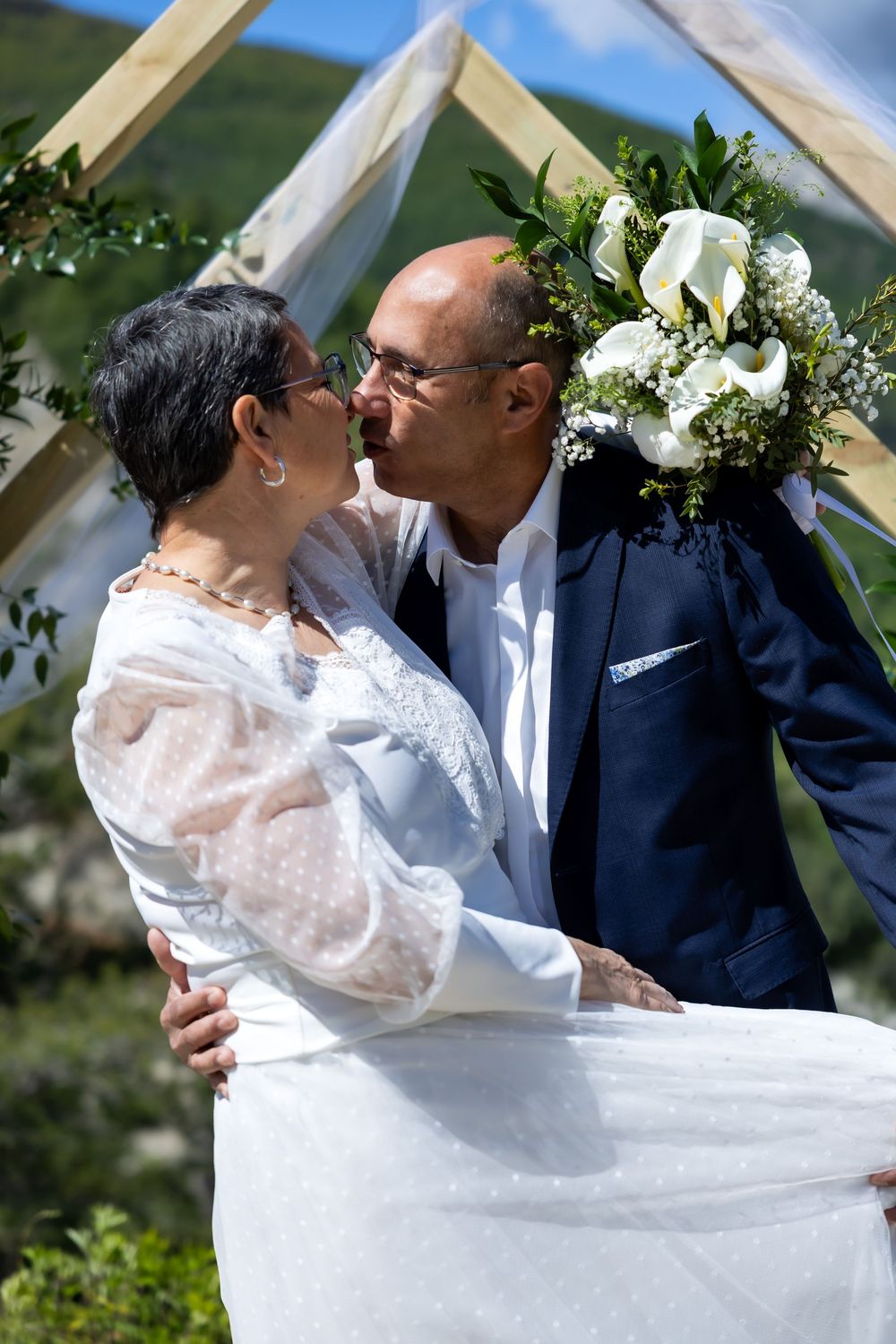 Mature couple sharing a kiss at outdoor wedding ceremony, bride in white dress holding floral bouquet under wooden arch.