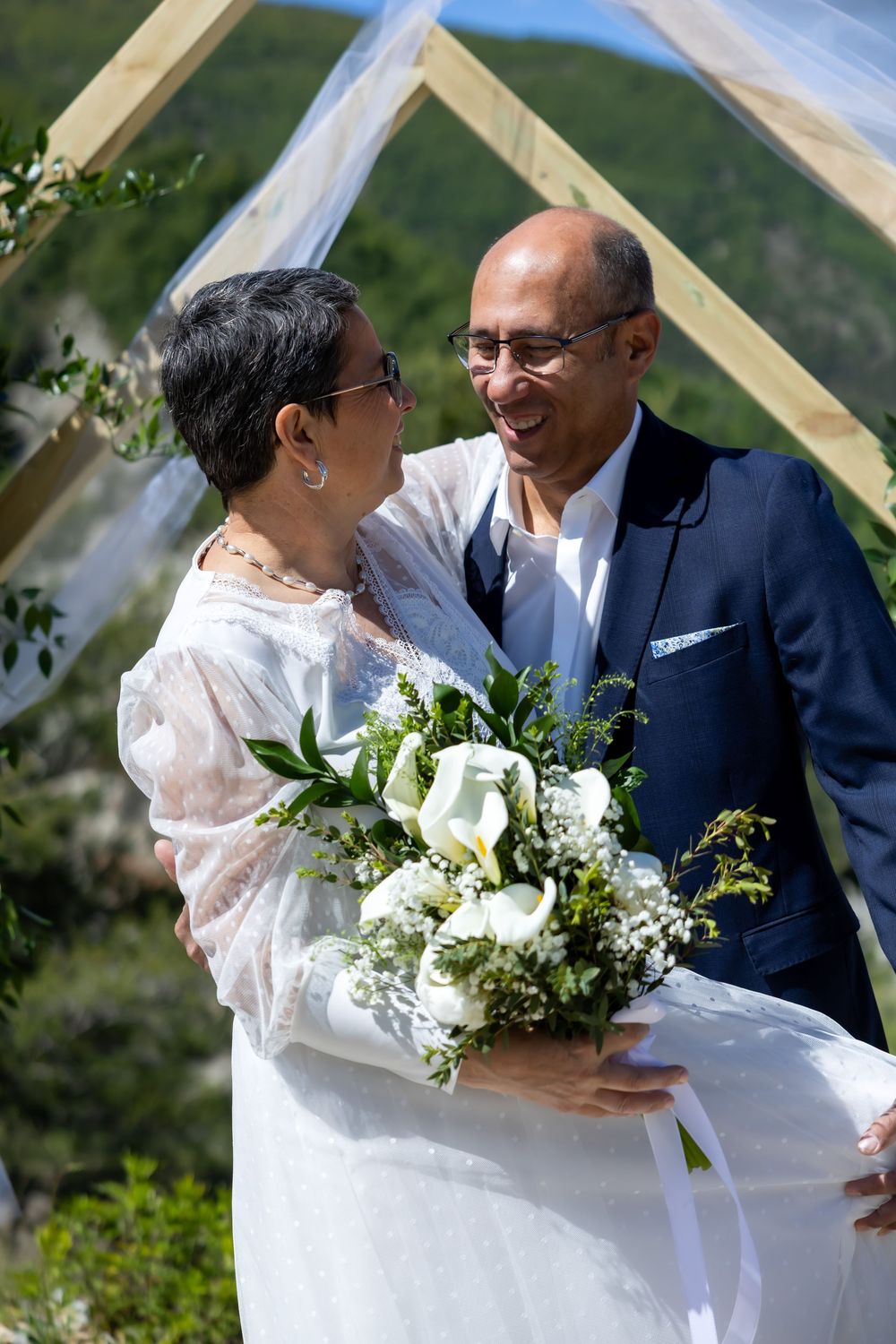 Mature couple in wedding attire smiling at each other outdoors, bride holding white calla lily bouquet under wooden arch.