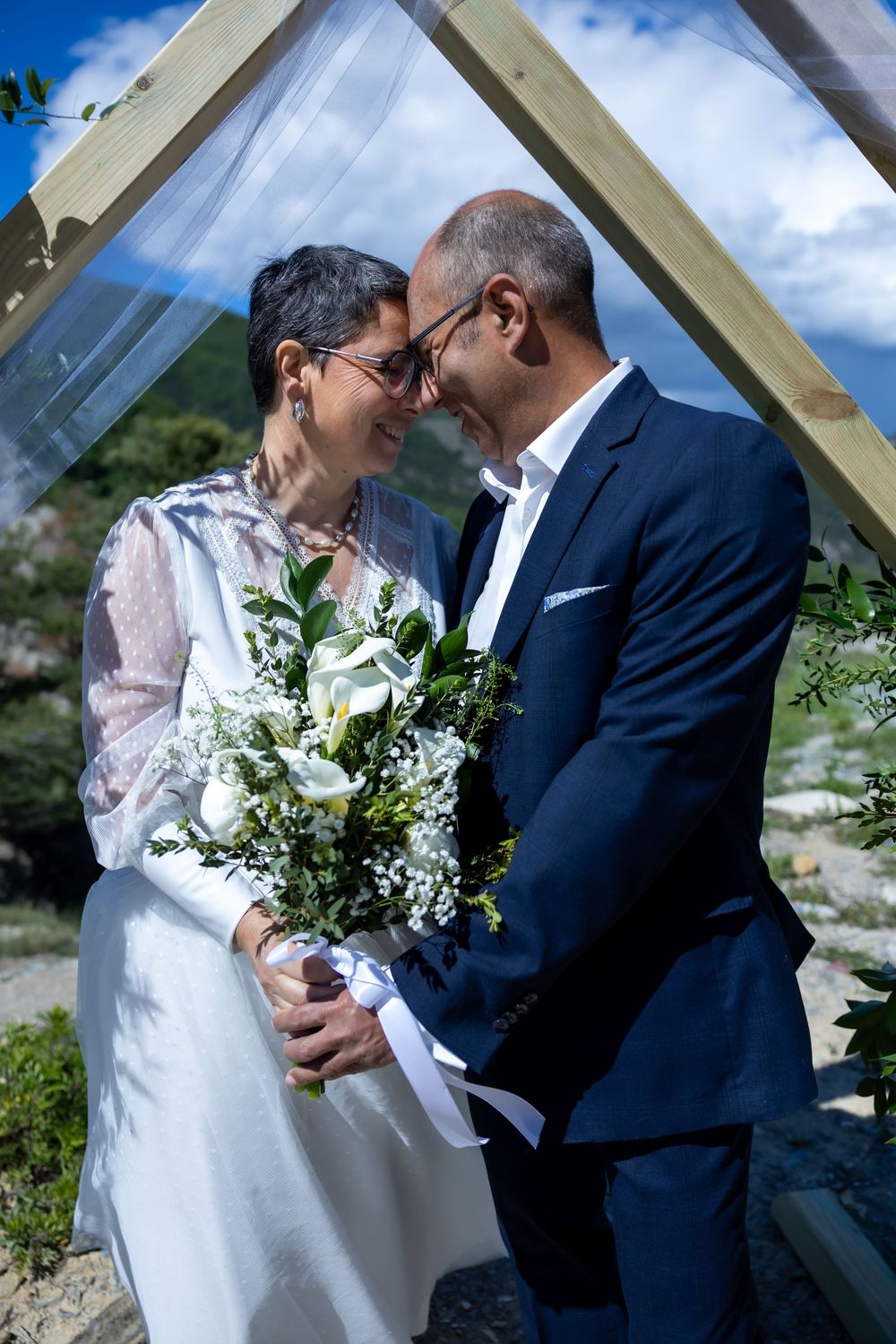 Couple in wedding attire sharing a romantic moment, bride holding white floral bouquet outdoors under wooden arch.