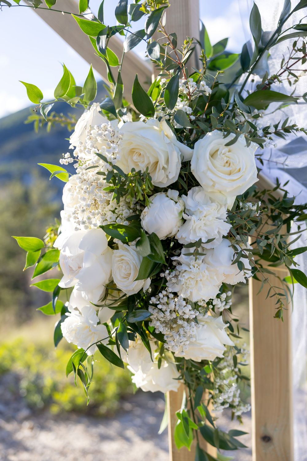 White roses and peonies floral arrangement on a wooden wedding arch with greenery and mountain backdrop.