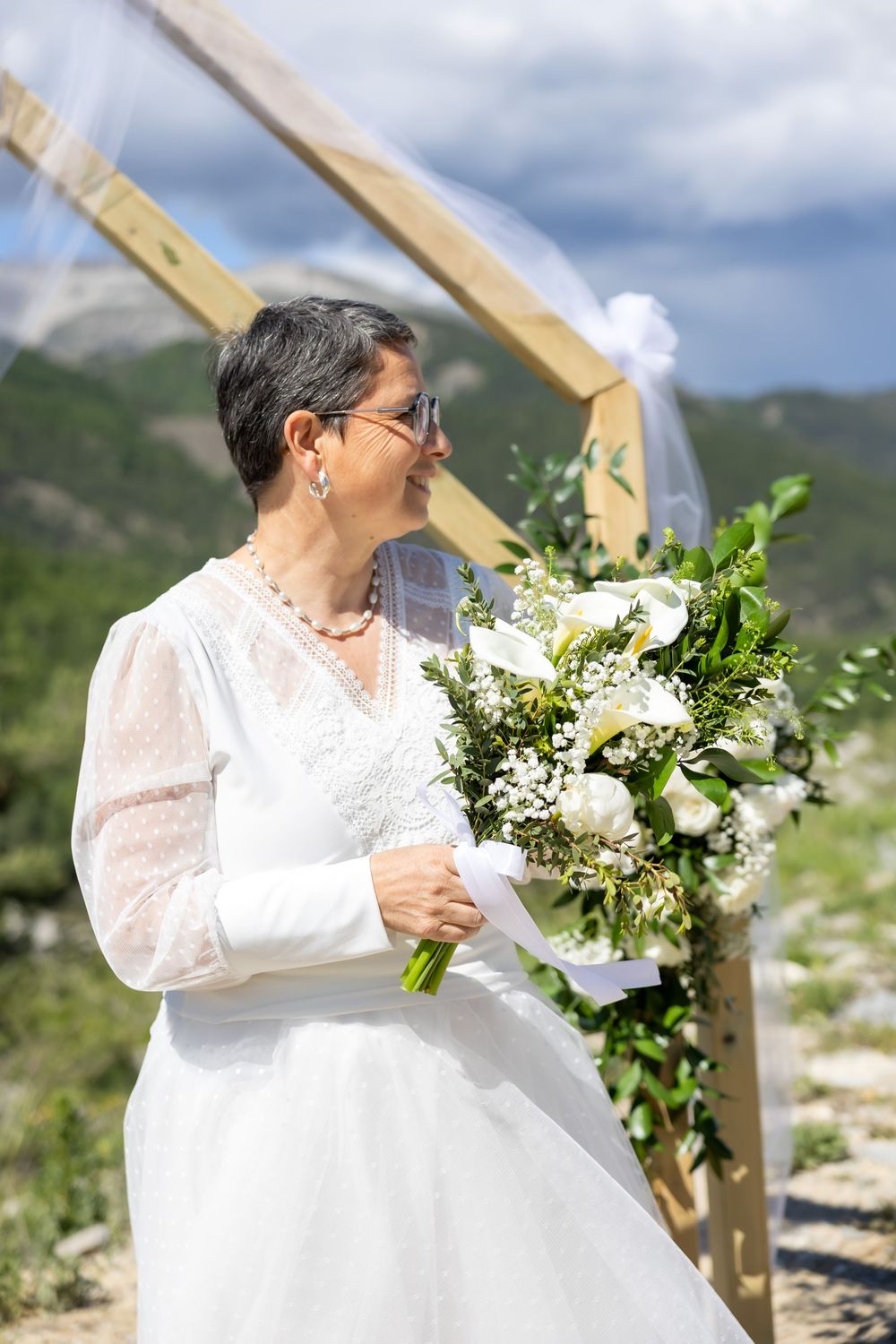 Mature bride in white dress holding bouquet of white flowers at outdoor mountain wedding ceremony.