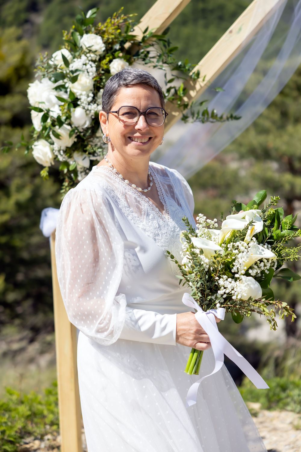 Smiling bride in white lace dress holding floral bouquet stands before wooden arch with white roses outdoors.