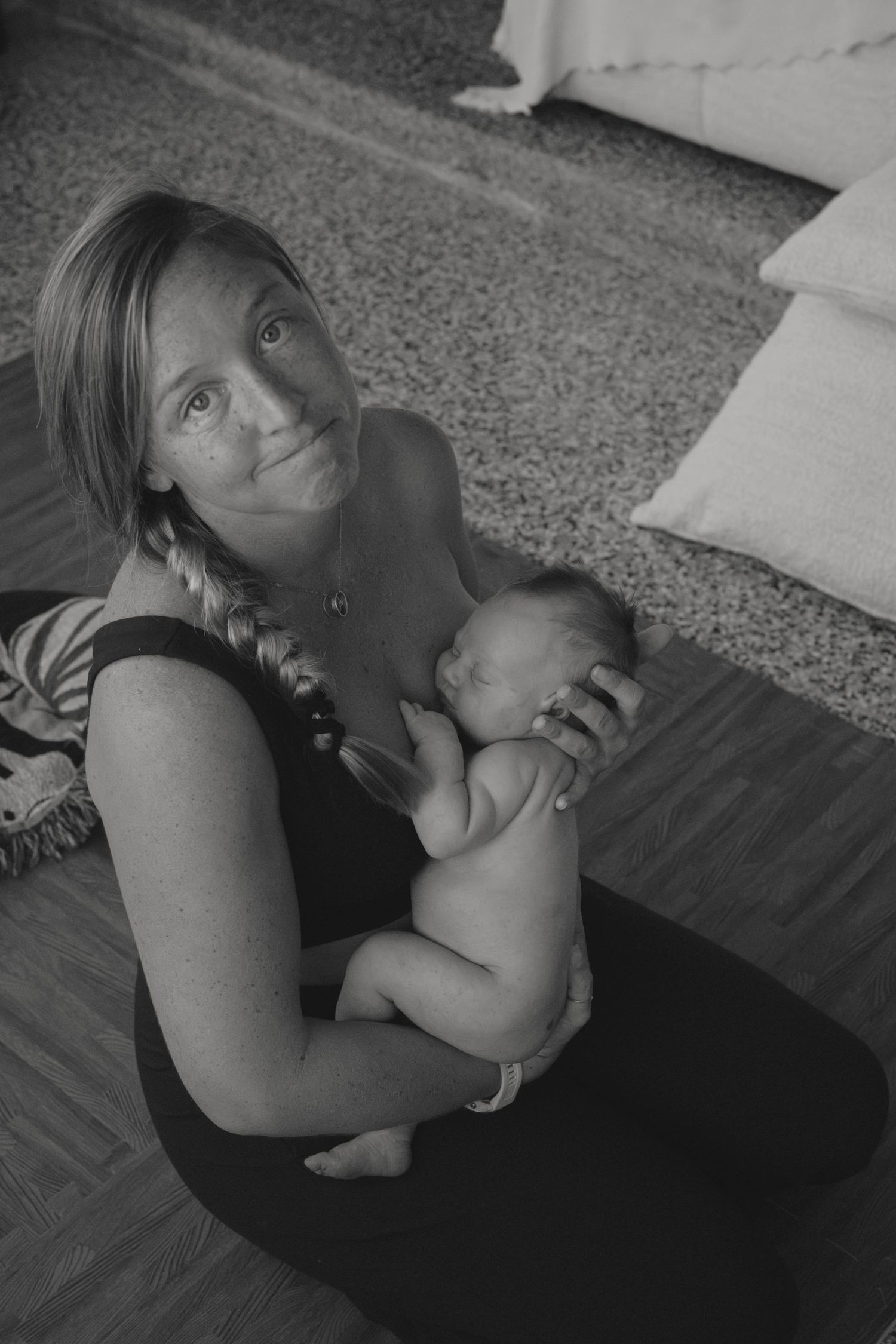 Black and white photo of a mother sitting on floor holding her newborn baby, looking up at camera with a gentle smile.