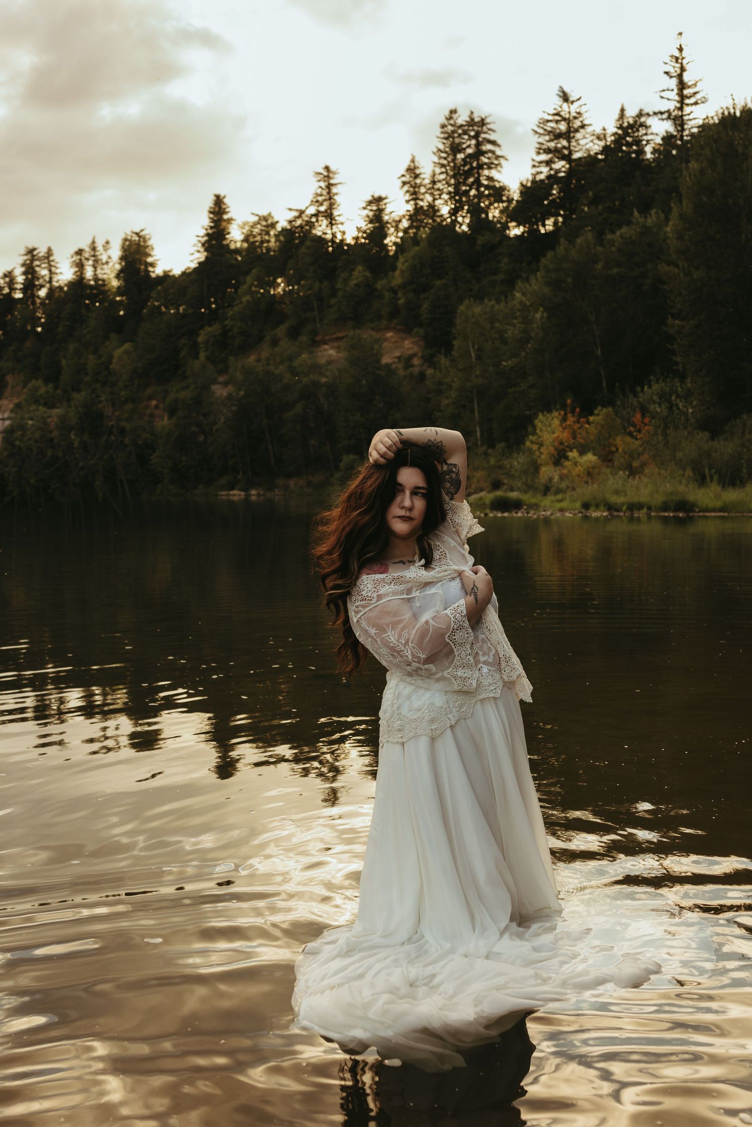 Woman in white lace dress standing in a lake with forested hills at golden hour.
