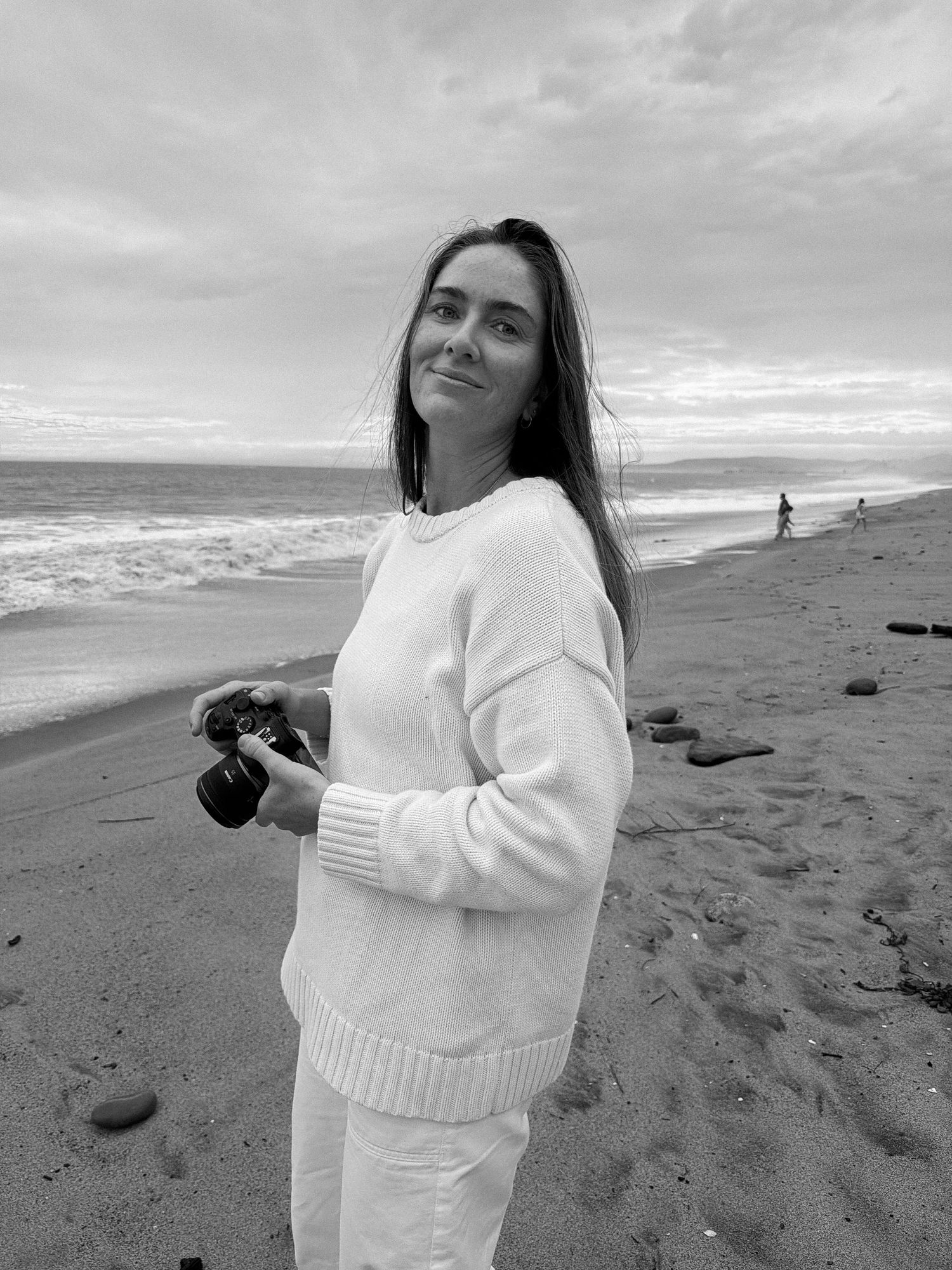 Woman in white sweater holding camera smiles on beach, ocean waves and cloudy sky in background, black and white photo.