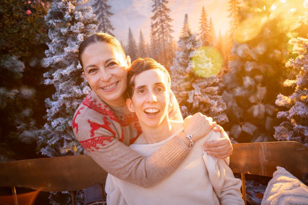 Two people smiling and hugging outdoors in a snowy winter setting with snow-covered trees and warm golden sunlight.