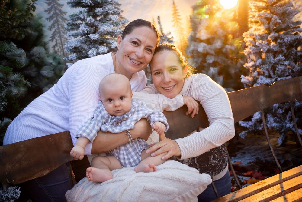 Two women and a baby pose together in front of snow-covered Christmas trees in a warm holiday photo session.