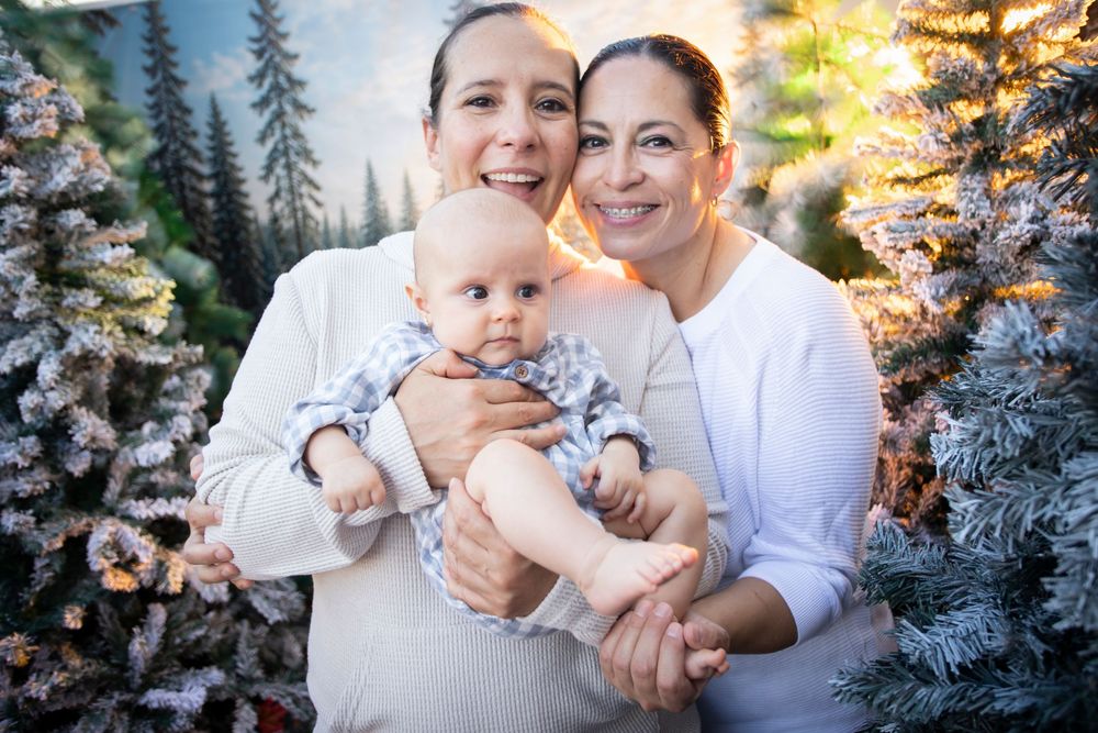 Two women in white tops smile warmly while holding a baby in a festive Christmas tree setting.