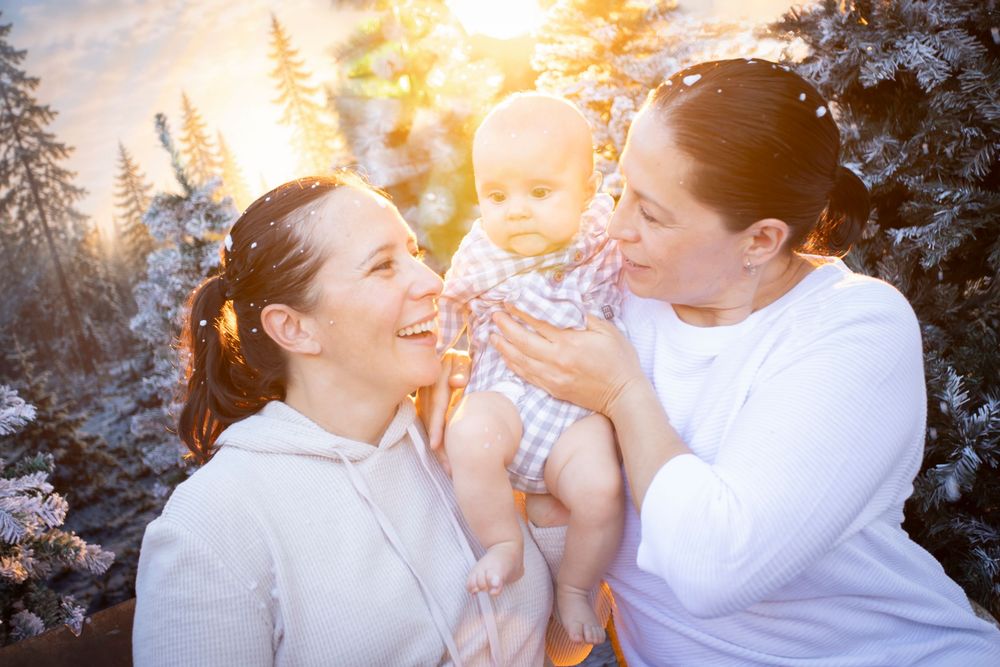 Two women smiling and holding a happy baby outdoors in a snowy winter forest with golden sunset light.