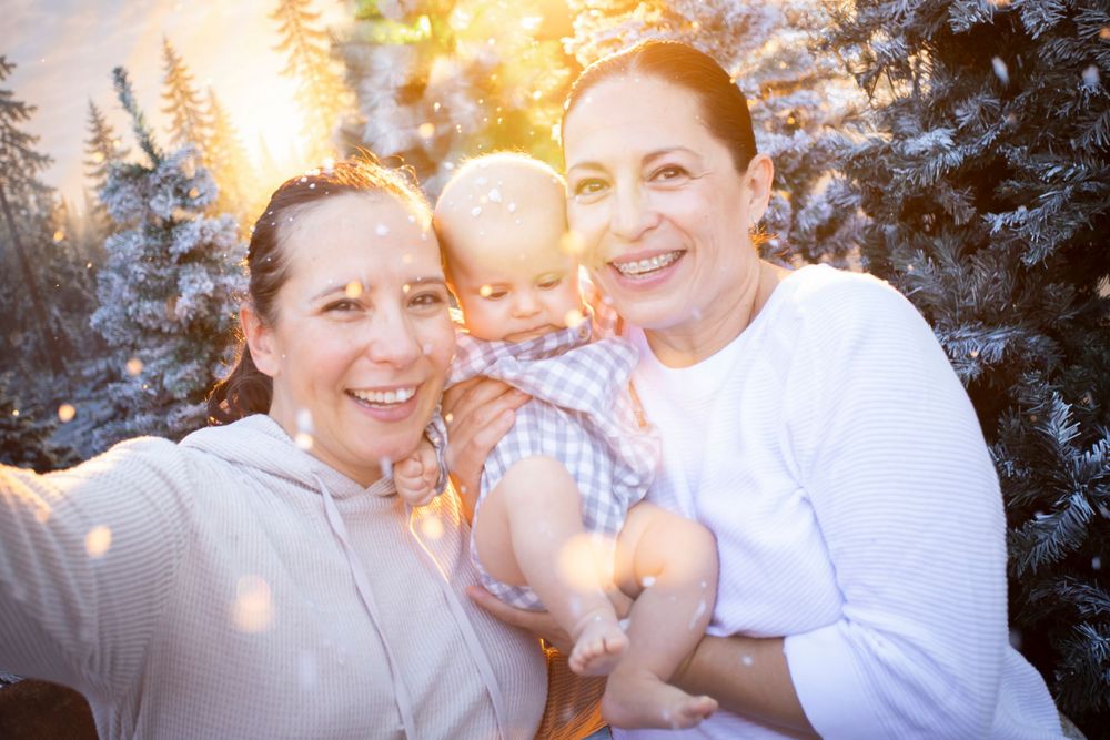 Two women and a baby smile together outdoors near snow-covered trees in warm winter light.