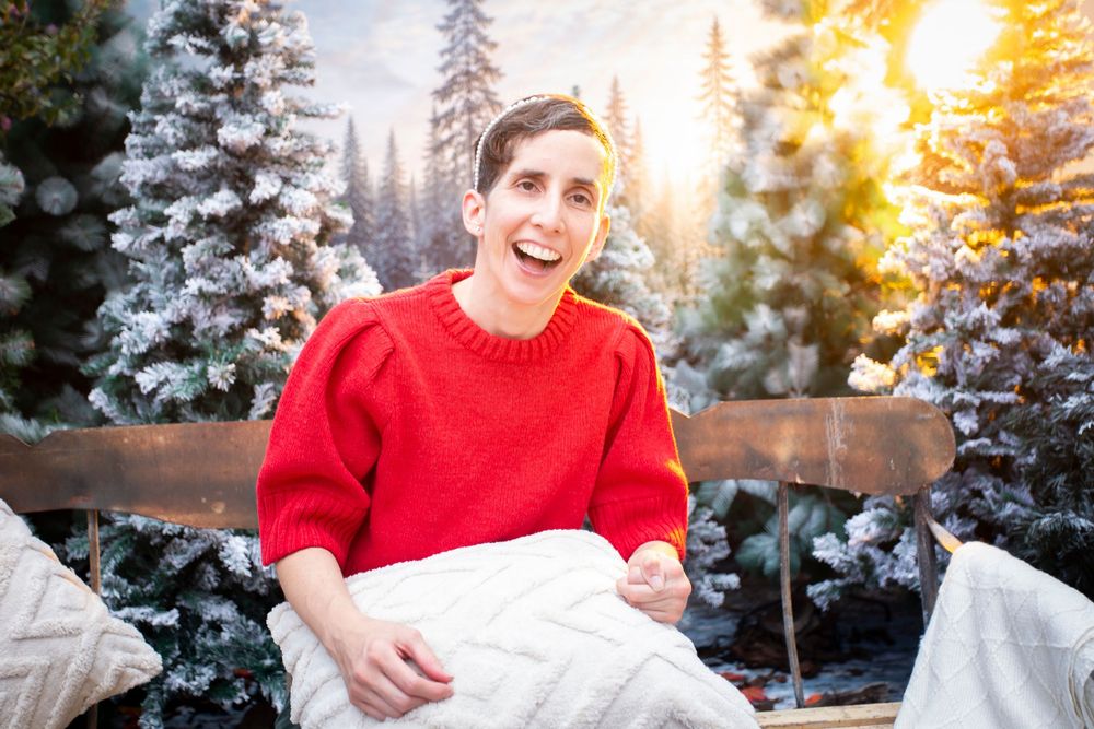 Person in red sweater laughing joyfully outdoors among snow-dusted pine trees with warm golden sunlight in background.