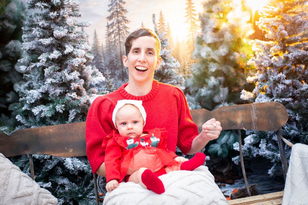 Man in red sweater holding baby in red outfit on bench with snowy Christmas trees backdrop.