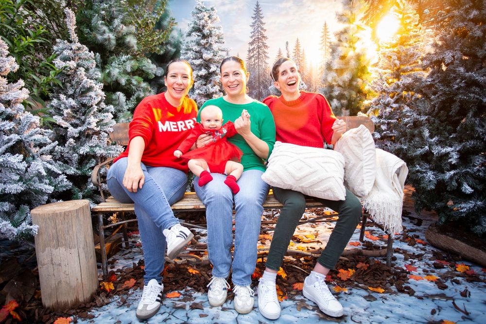 Three women in festive Christmas sweaters sit on a bench with a baby, surrounded by snow-covered trees.
