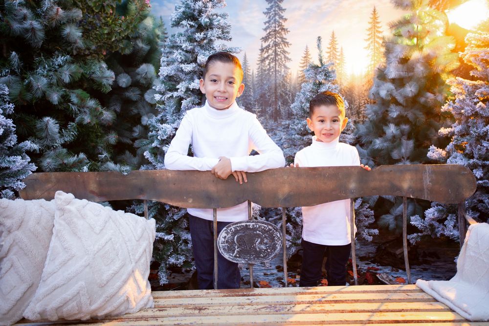 Two children in white tops pose by a wooden sled surrounded by snow-covered Christmas trees in a festive winter scene.