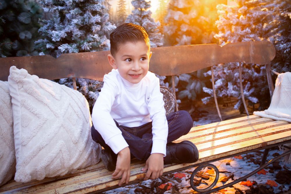 Young boy in white shirt sits cross-legged on wooden bench with pillows, surrounded by snow-dusted trees in warm sunset light.