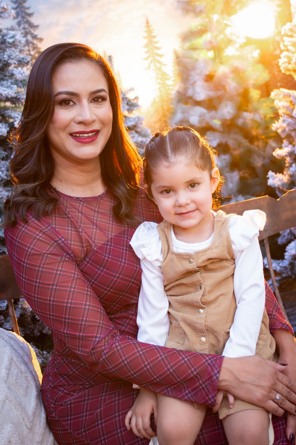 A mother in plaid dress holds her young daughter on a bench surrounded by snow-dusted Christmas trees in golden light.