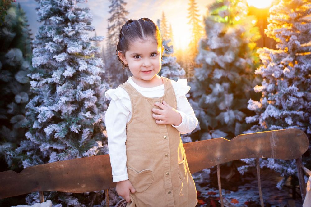 Young girl in tan jumper and white blouse poses by snowy pine trees in warm golden sunlight.