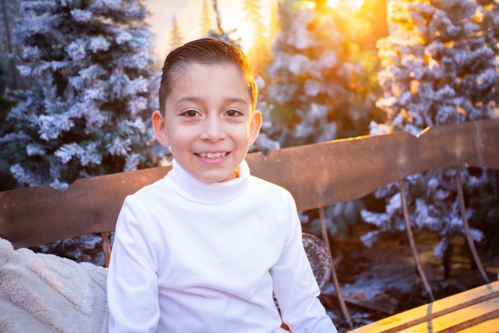 Smiling young boy in white turtleneck sitting outdoors in winter with snow-covered trees and golden sunset light.
