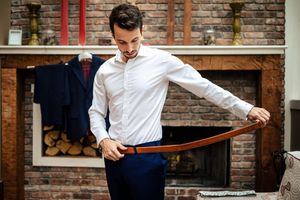 Man in white shirt fastening a brown leather belt over navy trousers near a brick fireplace.