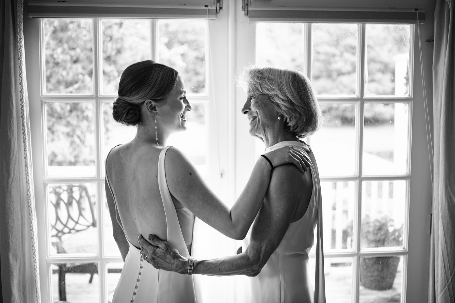 Black and white photo of a bride and her mother sharing a tender moment by a window on the wedding day.