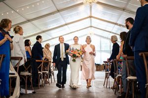 Bride walks down the aisle with parents in elegant tent wedding venue with string lights and colorful bouquet.
