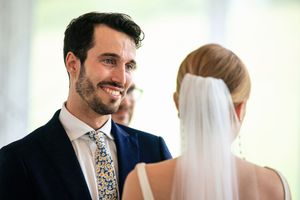 Groom in navy suit smiling at bride in veil during outdoor wedding ceremony.