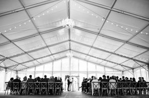 Elegant outdoor wedding ceremony under a large white tent with string lights and chandelier, guests seated in wooden chairs.