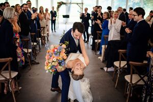 Groom dips bride in wedding aisle while guests applaud, bride holding colorful bouquet in elegant venue.