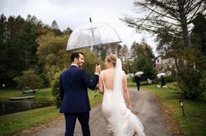 Bride and groom walking in rain under clear umbrella on lush green path in elegant wedding attire.