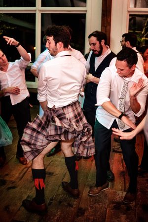 Men dancing energetically at a wedding reception, one wearing a traditional plaid kilt with red socks on a wooden dance floor.