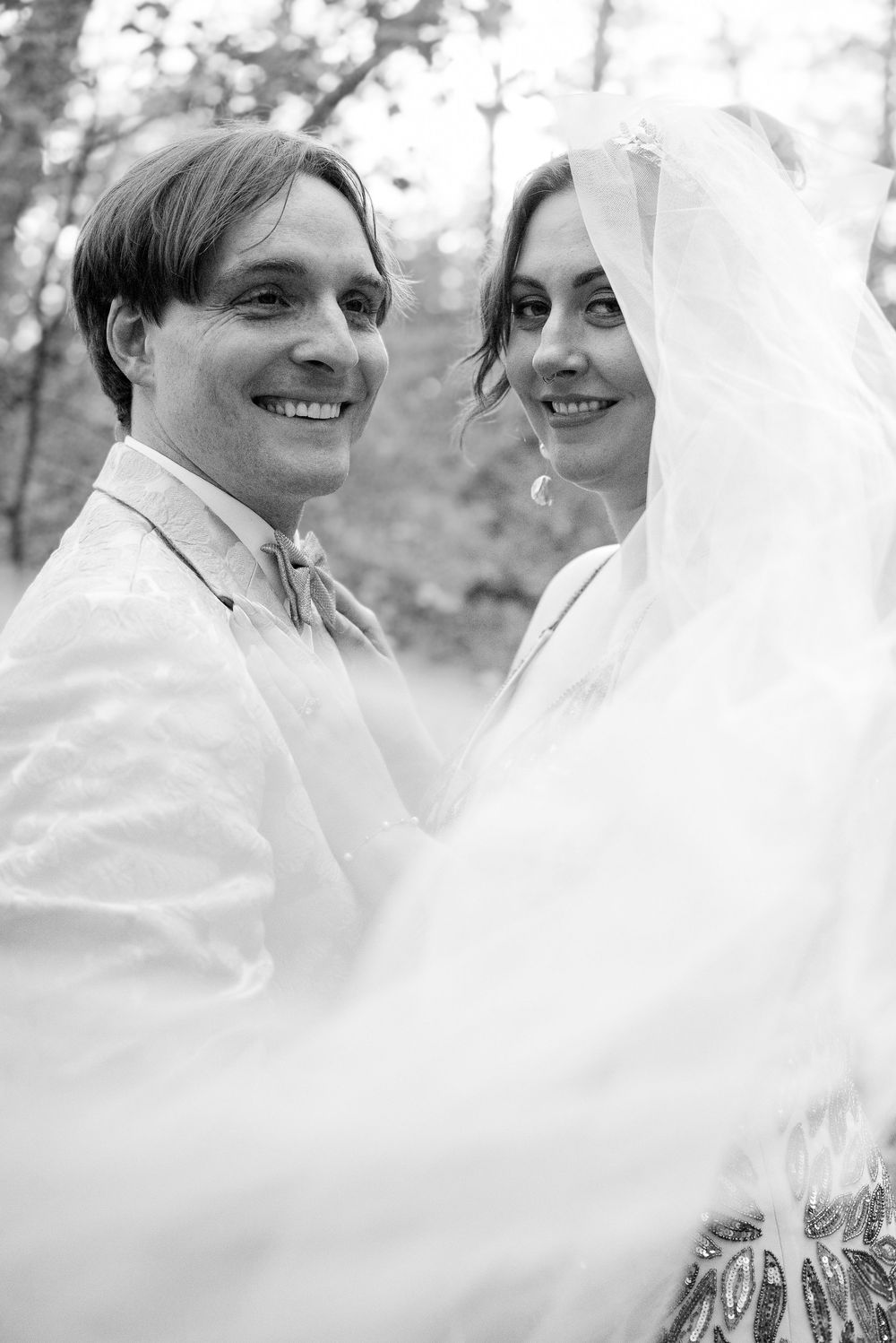 Black and white wedding portrait of smiling bride and groom outdoors, veil flowing in the breeze.