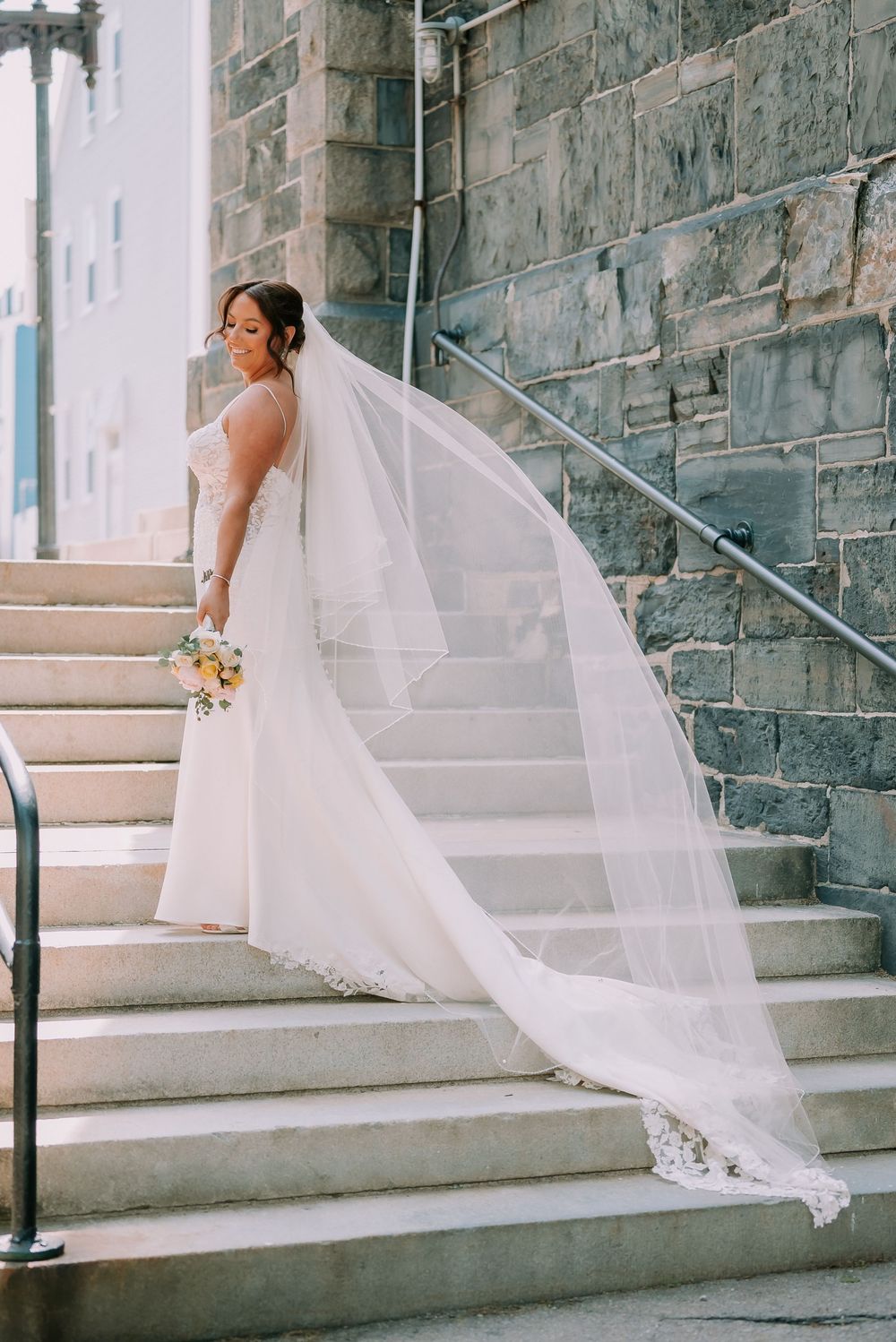 Bride in white gown with long lace veil flowing on stone church steps, holding yellow bouquet.