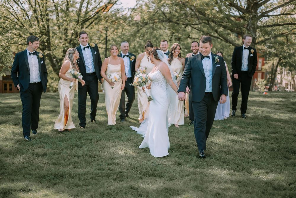 Wedding party walks outdoors on green lawn, bride and groom lead groomsmen in tuxedos and bridesmaids in champagne gowns.
