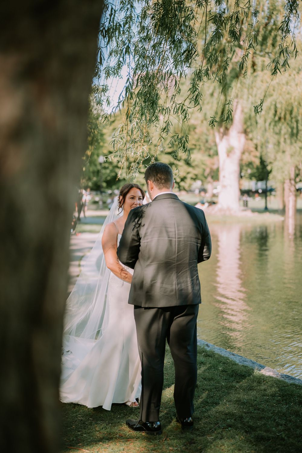 Bride and groom share a tender moment by a serene pond under a weeping willow tree during their wedding day.
