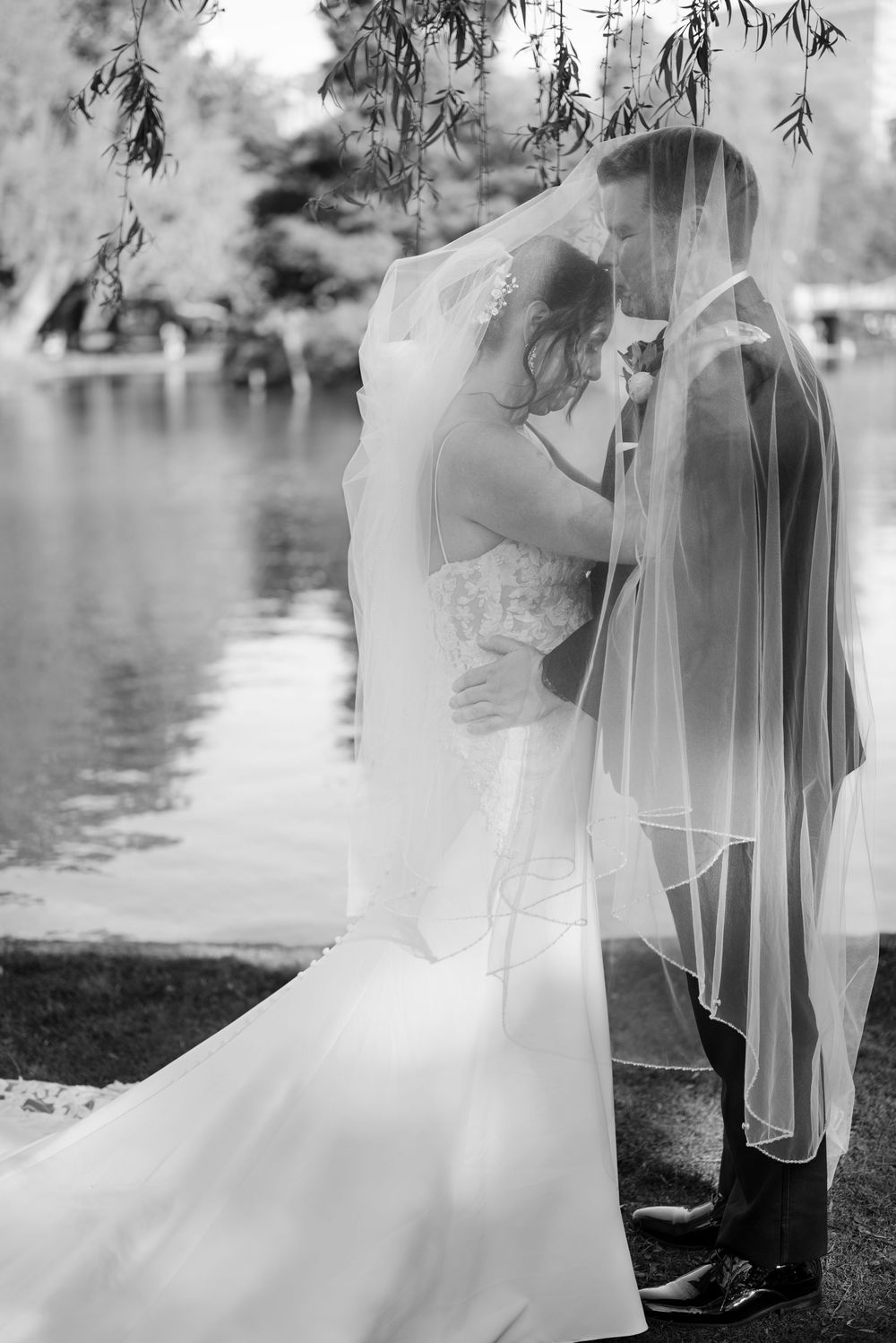 Black and white photo of bride and groom kissing under veil by a lake.