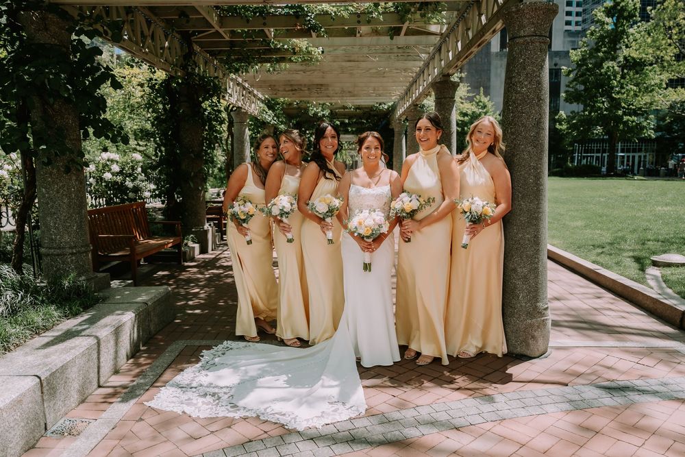 Bride in white gown with bridesmaids in yellow dresses holding bouquets under pergola in garden.