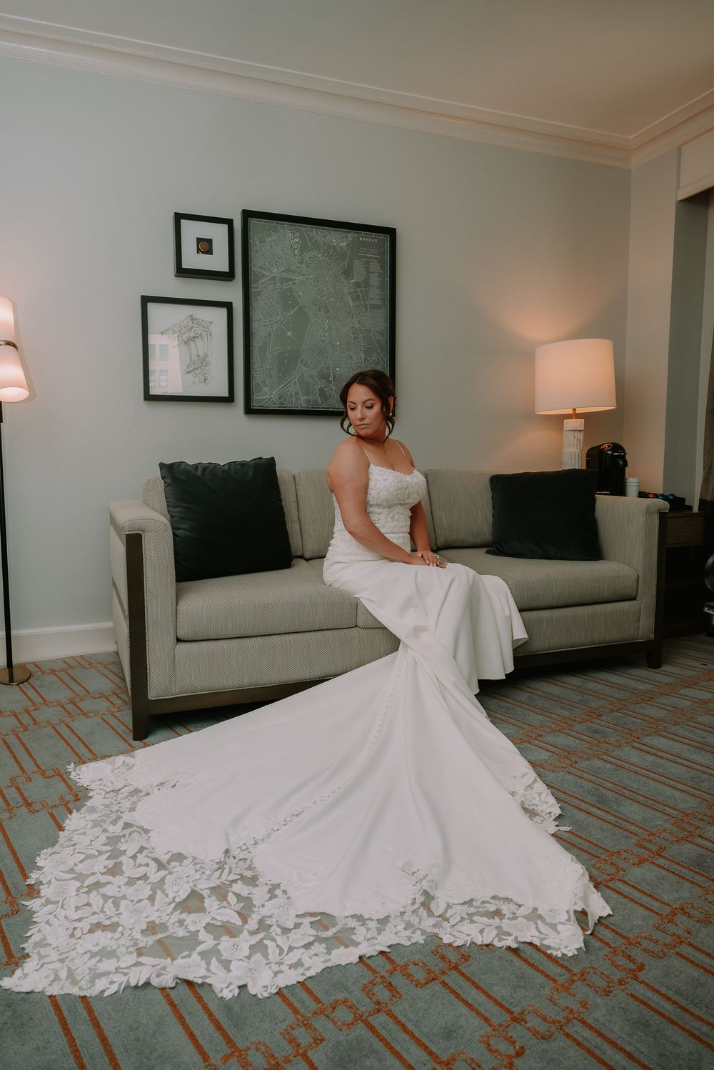 Bride in a white mermaid lace wedding gown with a long train seated elegantly on a sofa in a hotel suite.