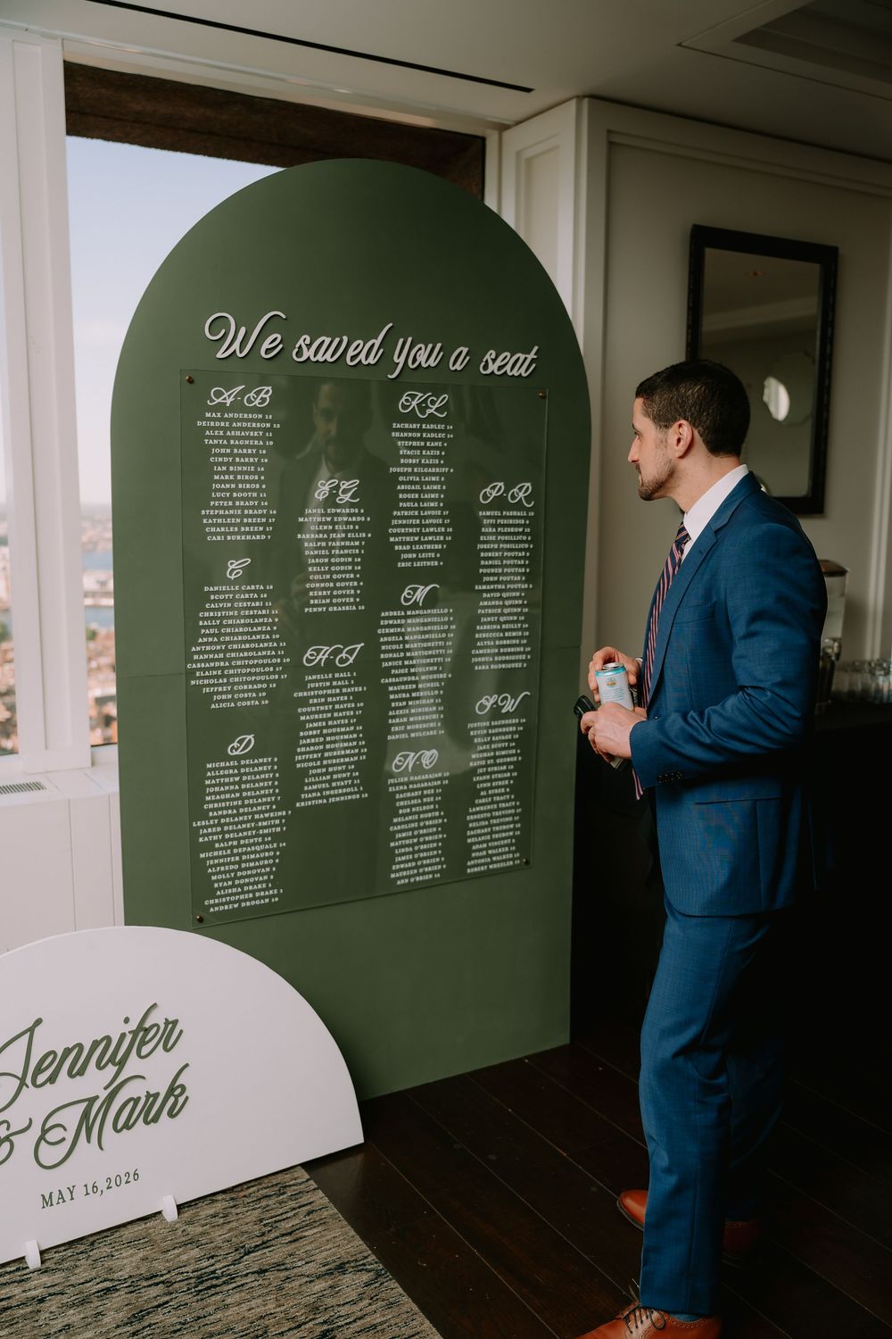 Groom in blue suit reviews wedding seating chart on green arched sign reading We Saved You a Seat.