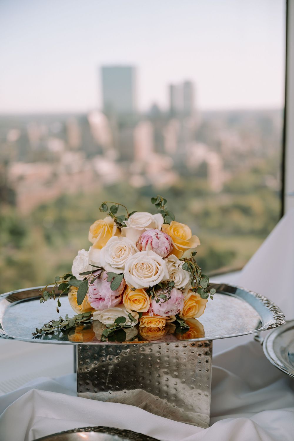 Elegant wedding bouquet of yellow, pink, and white roses on a silver tray with a city skyline backdrop.