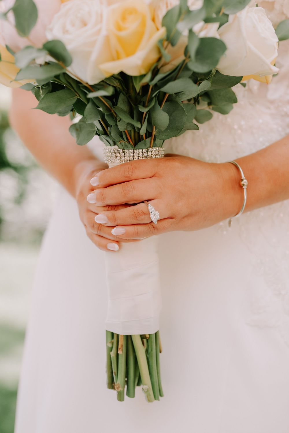 Close-up of bride holding yellow and white rose bouquet with diamond engagement ring and pearl bracelet.