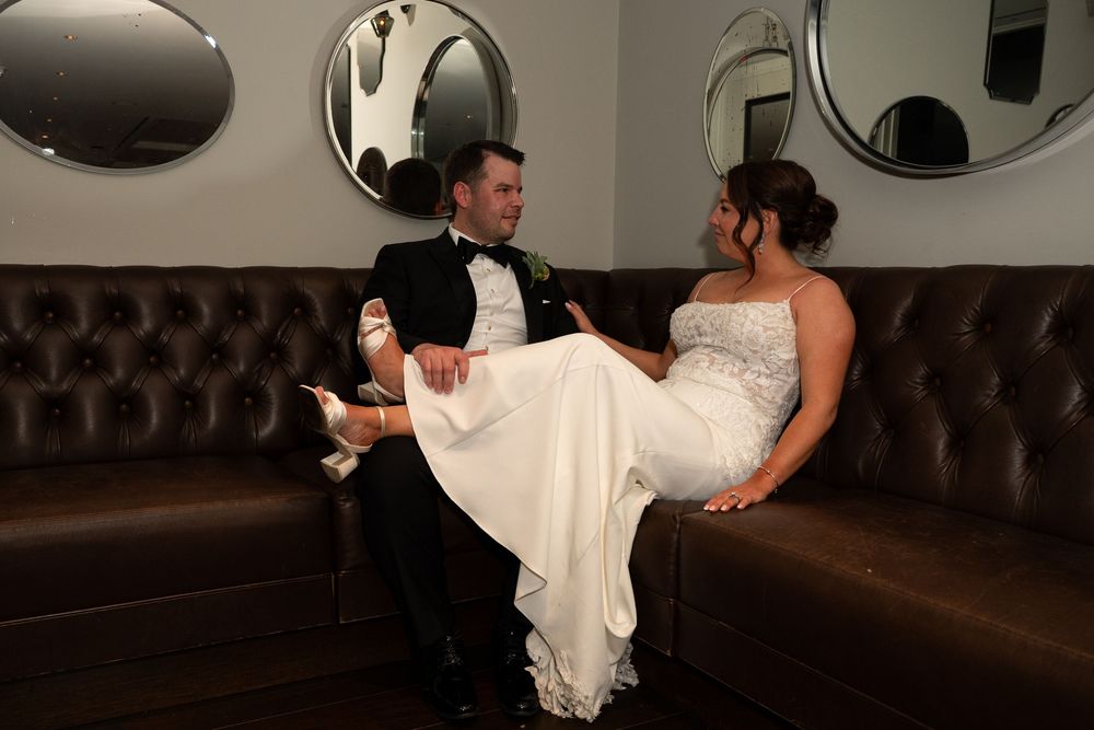 Bride and groom sitting together on a brown leather sofa, smiling, with the bride in a white gown and groom in a tuxedo.